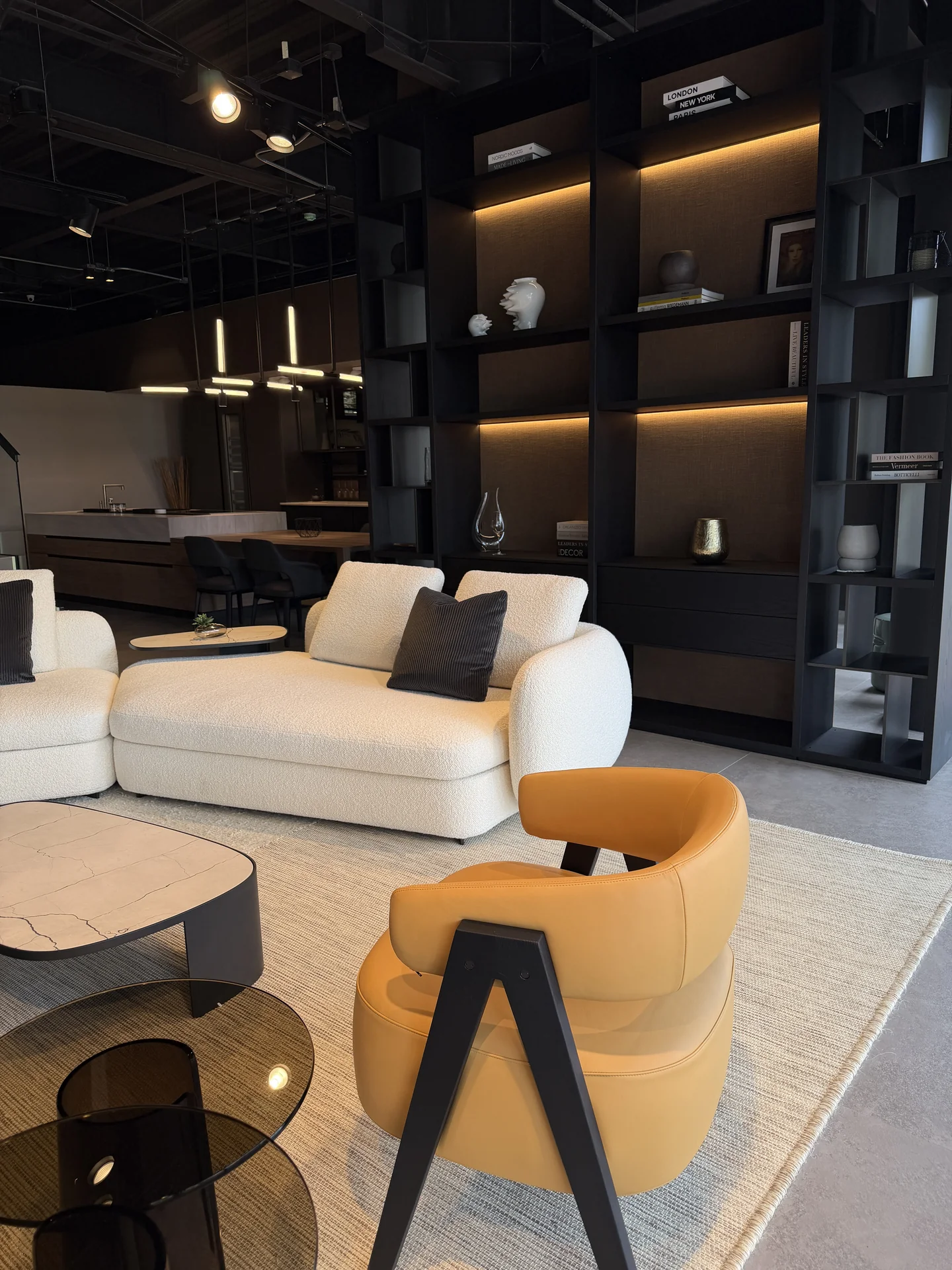 Sala Poliform con sofá modular en boucle blanco, sillón mostaza y librería de pared retroiluminada en madera oscura · showroom Productos Arquitectónicos en Bogotá